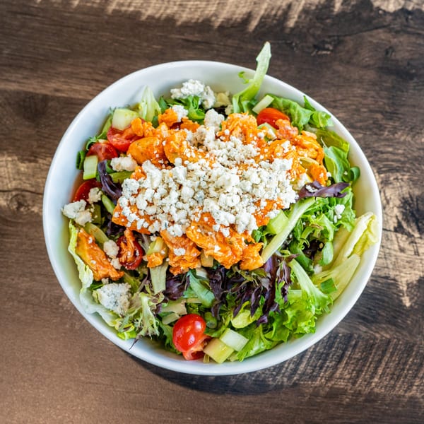 A vibrant Invictus Pizza Kitchen buffalo chicken salad with blue cheese crumbles in a white bowl.