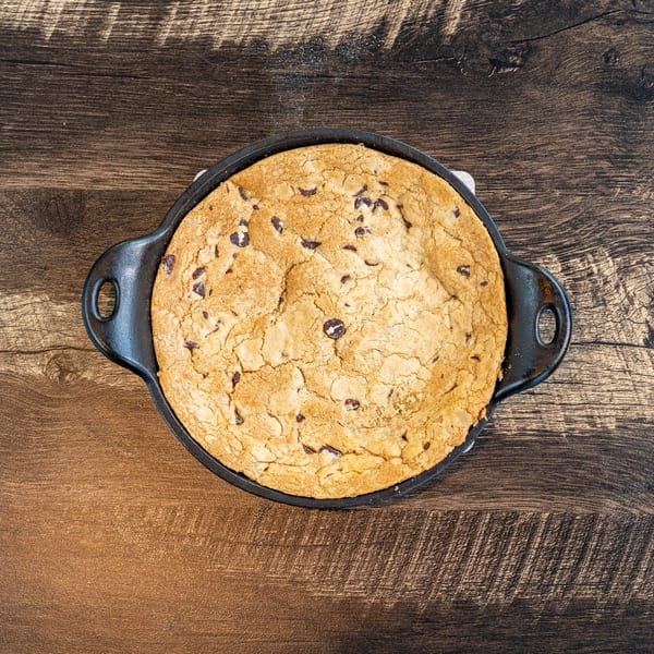 A freshly baked chocolate chip cookie in a cast iron skillet on a wooden table.